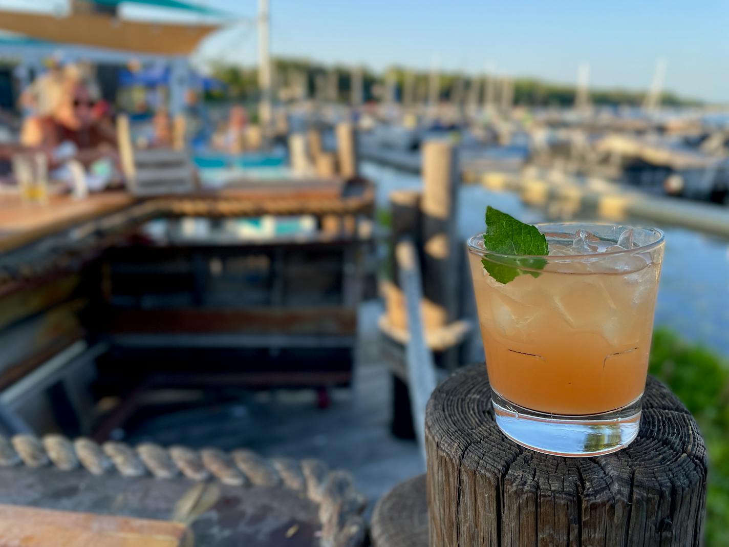 An orange cocktail with a mint leaf garnish on a wooden pole wrapped in thick rope by a bar shaped like a boat bow. The marina is visible in the background.