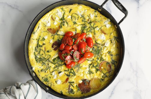 A skillet with an herb-filled potato, summer squash and goat cheese frittata with a cherry tomato sauce and fresh herbs on top.