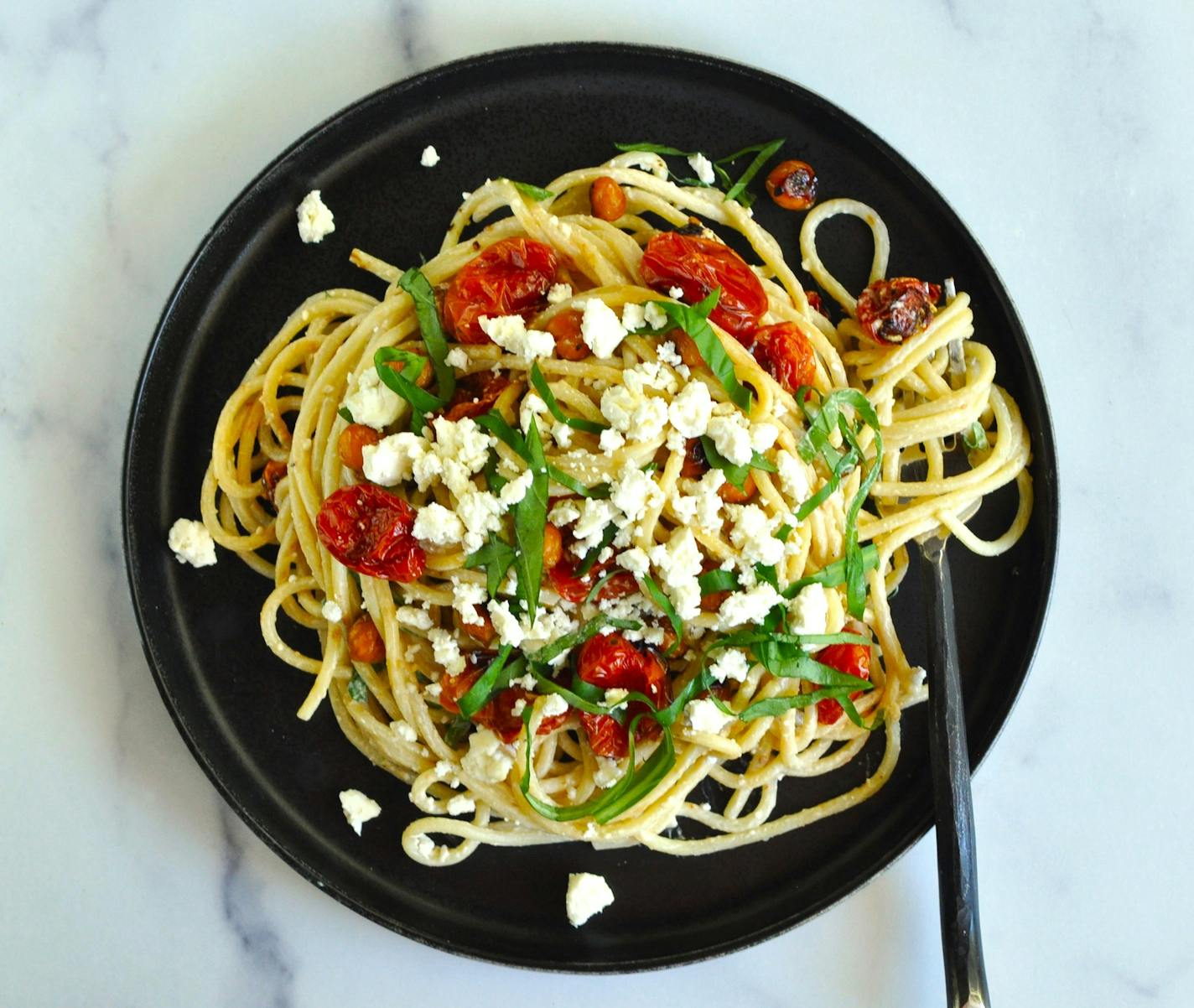 A bowl of pasta salad made with spaghetti noodles, roasted cherry tomatoes and chickpeas, flavored with a tahini dressing.