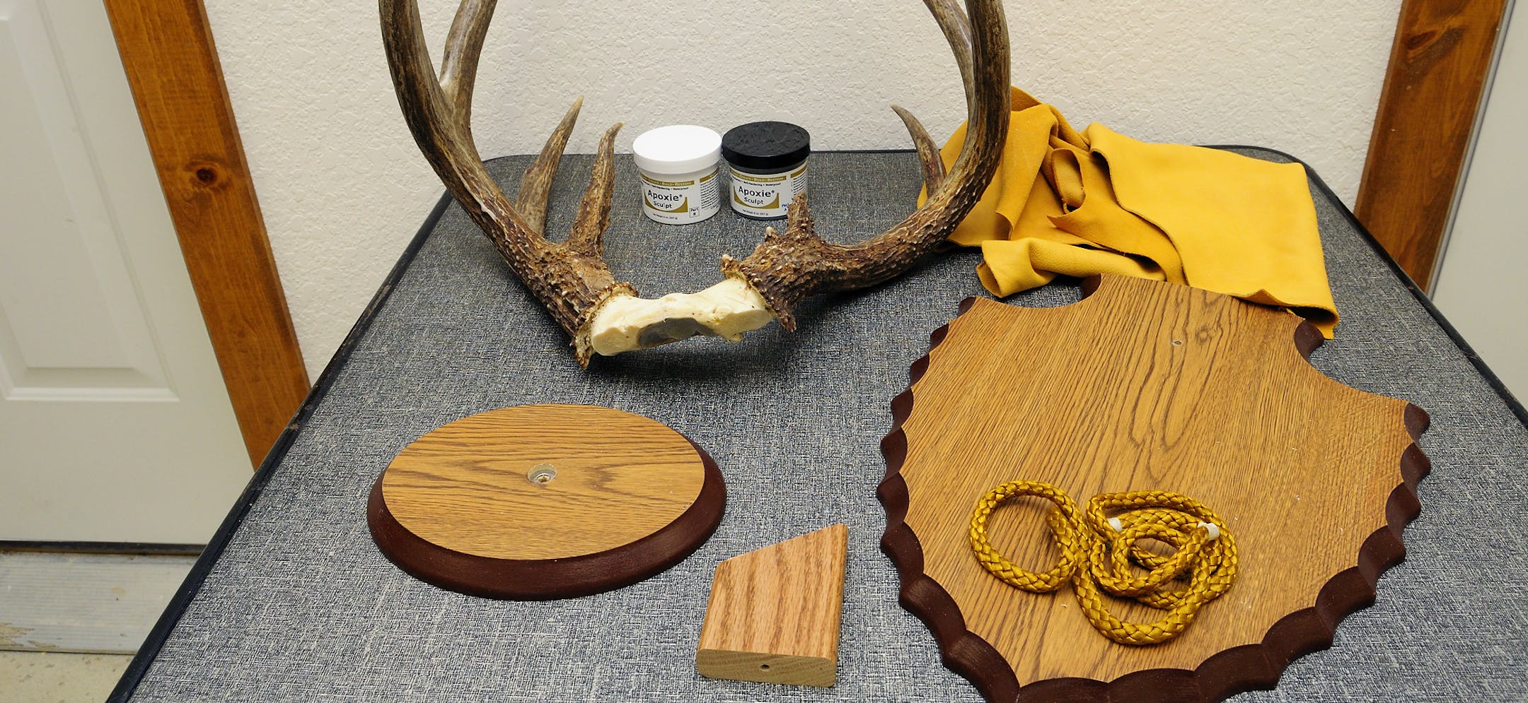 From top right, the materials needed for an antler plaque diplay include leather for covering the skull plate, a plaque, braided rope, angle board, small plaque for the base of the antlers, antlers with skull plate cleaned and dry, and epoxy putty.