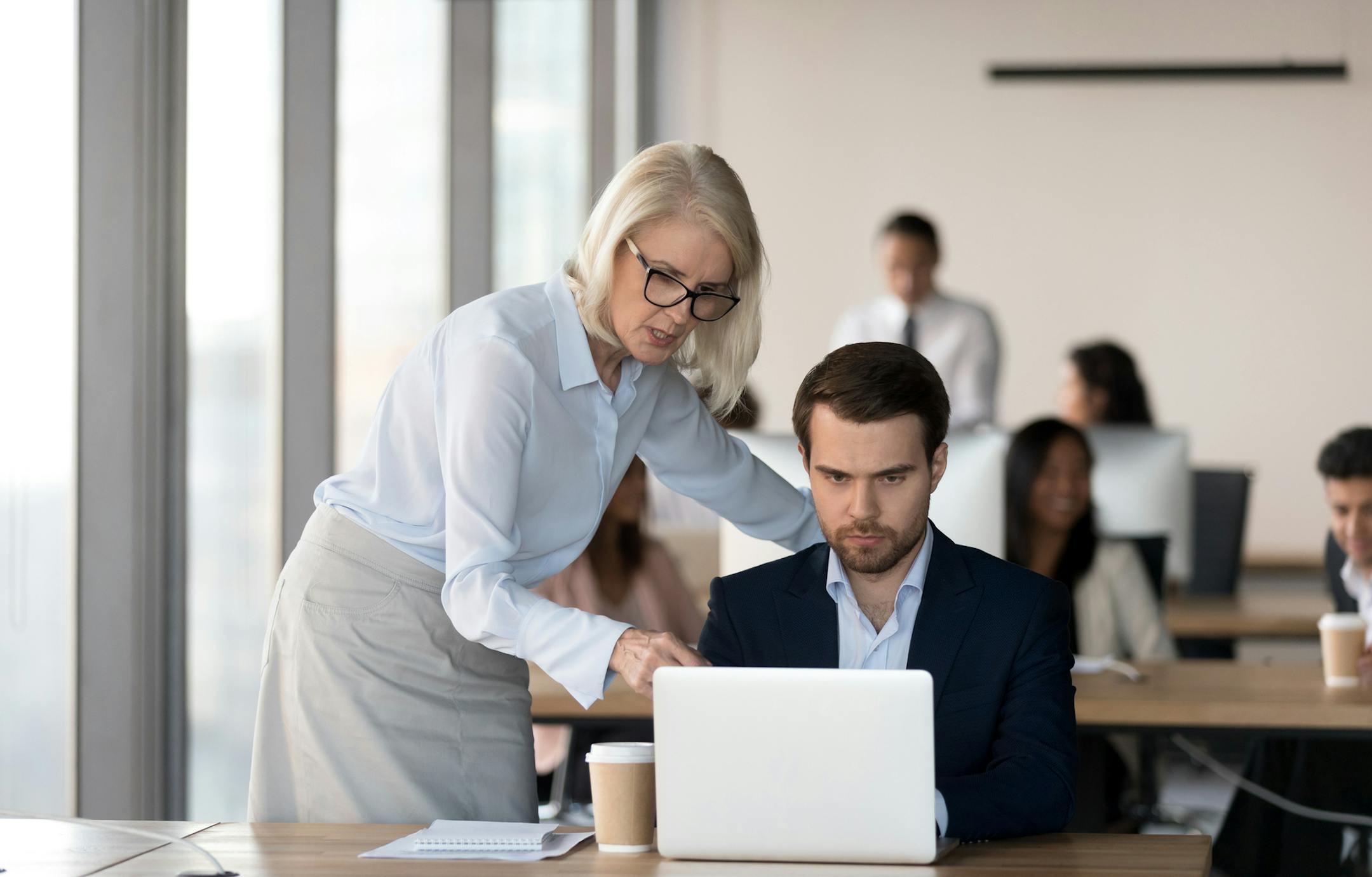 Middle aged female executive manager and male colleague working together in coworking space, businesswoman pointing on computer screen analyzing explaining business project results and discussing data