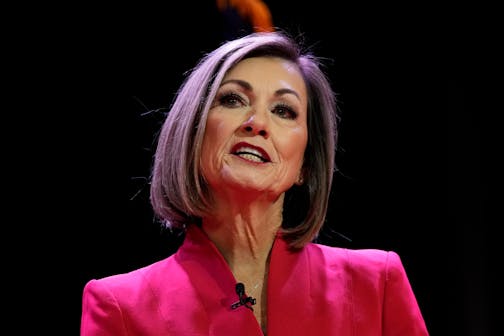 Iowa Gov. Kim Reynolds speaks against a dark background while wearing a dark pink suit.