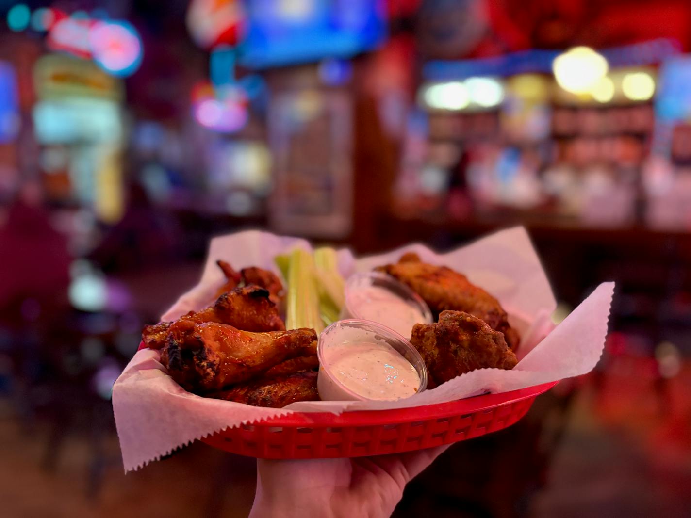 A red plastic basket holds two types of wings with a cup of ranch dividing them down the middle. In the background is an eclectic old bar with vintage neon signs and decor.