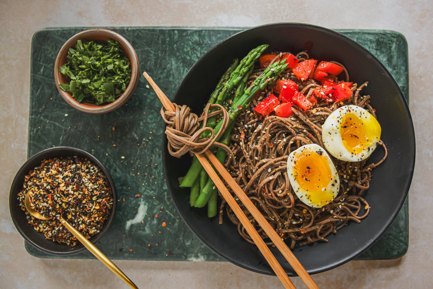 A bowl of soba noodle Bowl with Asparagus and Jammy and topped with shichimi togarashi, with small bowls of extra seasoning and herbs on the side.