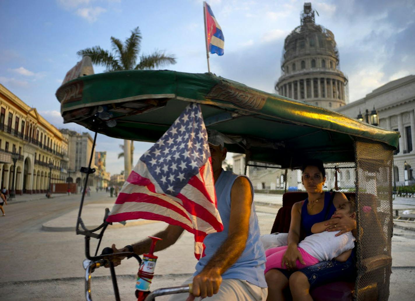 A taxi pedals his bicycle, decorated with Cuban and U.S. flags, as he transports a woman holding a sleeping girl, near the Capitolio in Havana, Cuba, Tuesday, March 15, 2016. President Barack Obama will travel to Cuba on March 20. (AP Photo/Ramon Espinosa)