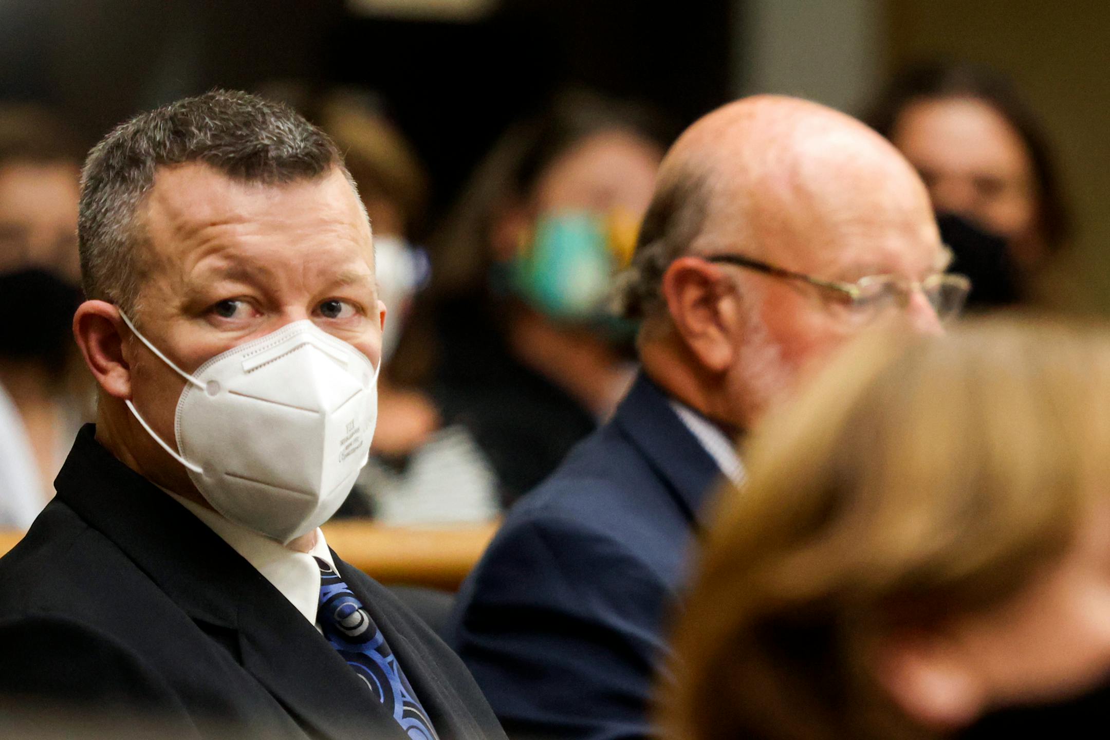 Paul Flores in dark suit and wearing face mask sitting next to male lawyer in suit in court.