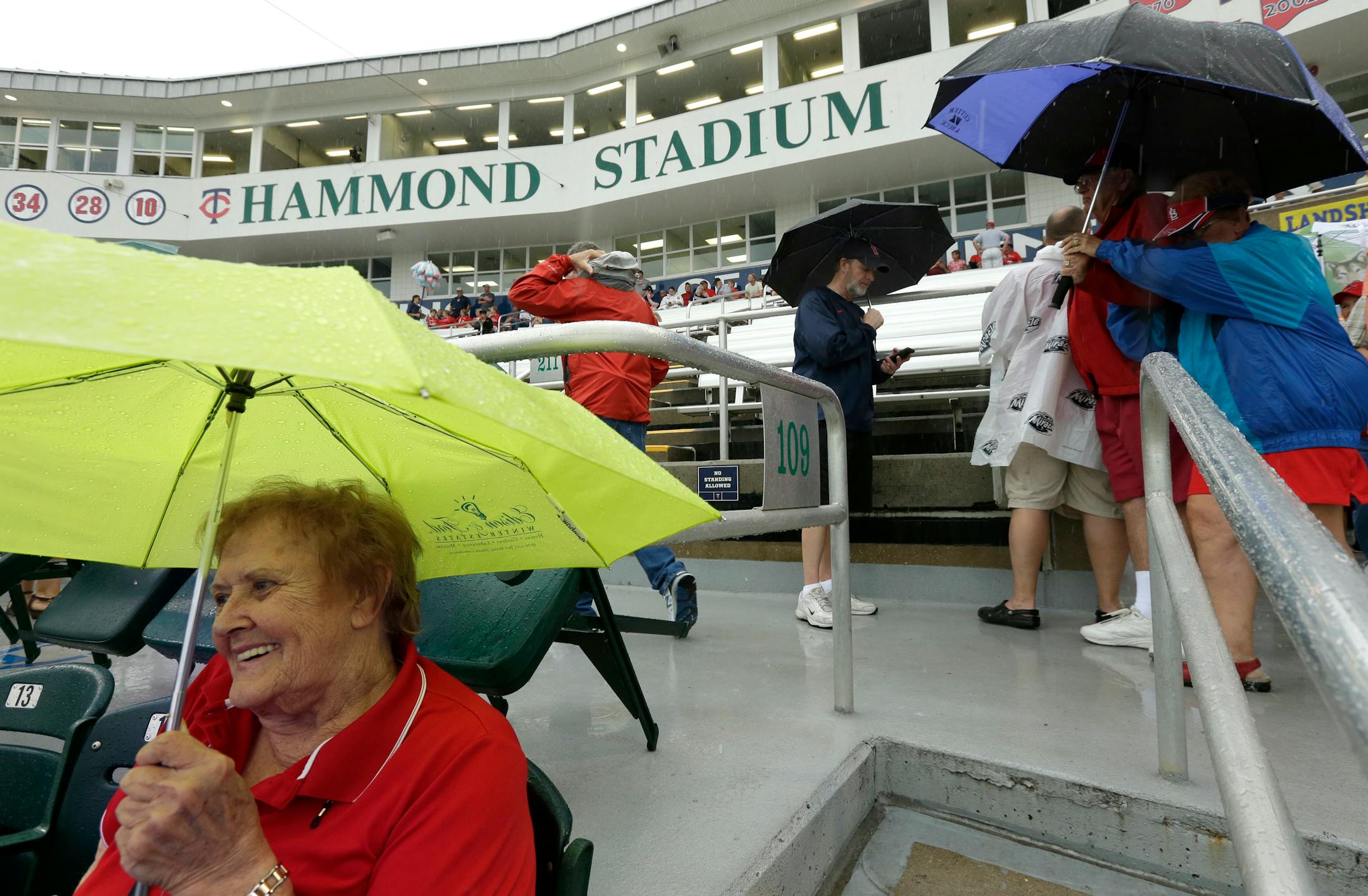 St. Louis Cardinals fan Alice Odenwald, of St. Louis, left, shelters herself from rain with an umbrella while sitting in the stands before a scheduled exhibition baseball game between the Cardinals and the Minnesota Twins, Thursday, March 6, 2014, at Hammond Stadium, in Fort Myers, Fla. (AP Photo/Steven Senne)