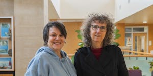 Two women smile and stand beside each other in a campus building.