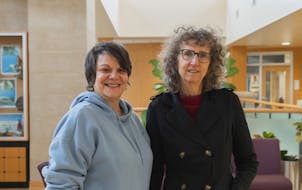 Two women smile and stand beside each other in a campus building.