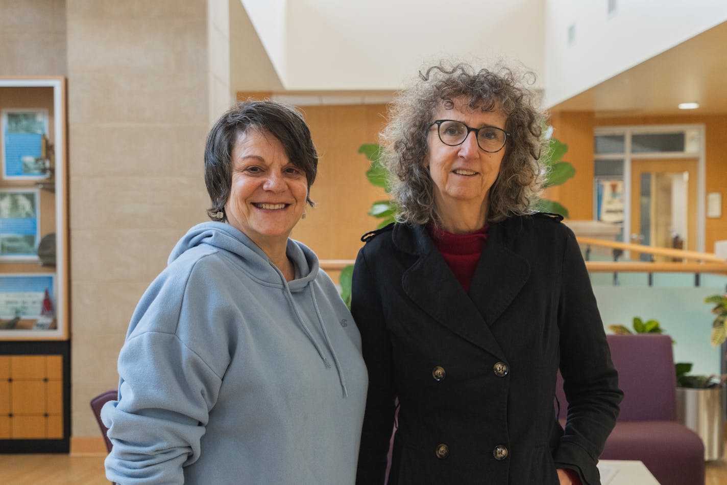 Two women smile and stand beside each other in a campus building.