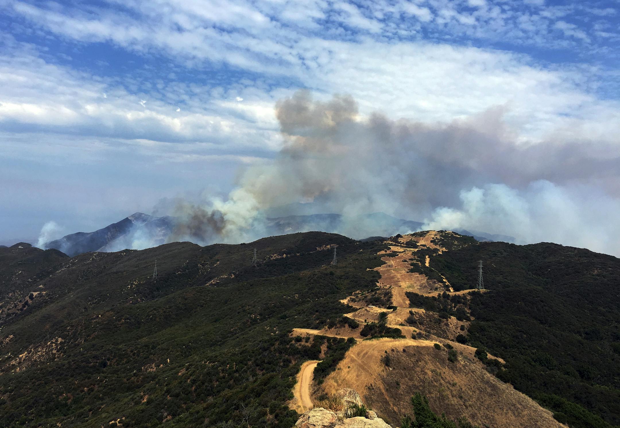 This photo provided by KEYT-TV shows smoke looming above Broadcast Peak behind a fire break along a ridge line east of Cachuma Lake in Santa Barbara County, Calif., Sunday, July 9, 2017. Wildfires barreled across the baking landscape of the western U.S. and Canada, destroying a smattering of homes, forcing thousands to flee and temporarily trapping children and counselors at a California campground. Southern California crews hope slightly cooler temperatures and diminishing winds will help in th