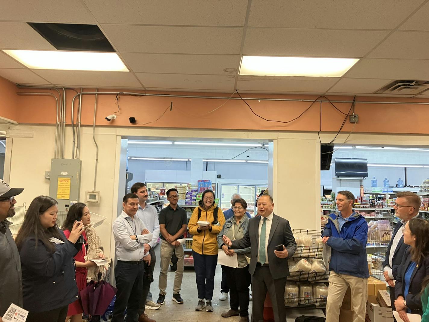 A man in a gray suit speaks and gestures as he stands in the middle of a circle of people in the front of a small grocery store.