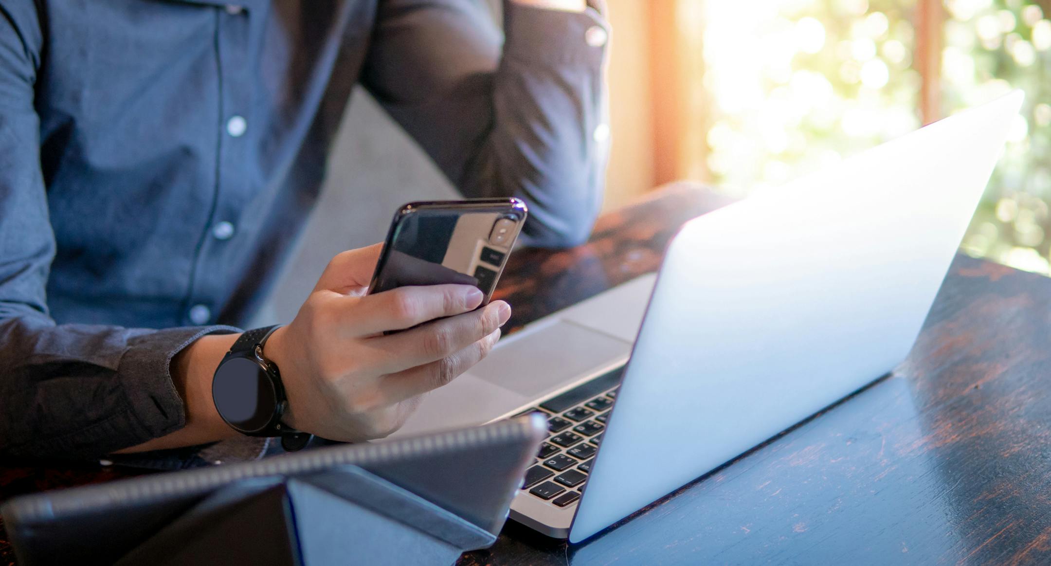 Male hand holding smartphone. Businessman using laptop computer and digital tablet while working in the cafe. Mobile app or internet of things concepts. Modern lifestyle in digital age.