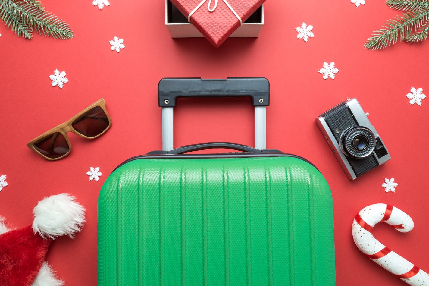 Christmas holiday minimal creative composition made of suitcase, gift box, sunglasses,Santa hat, candy cane, retro photo camera, fir branches and snowflakes on red background.