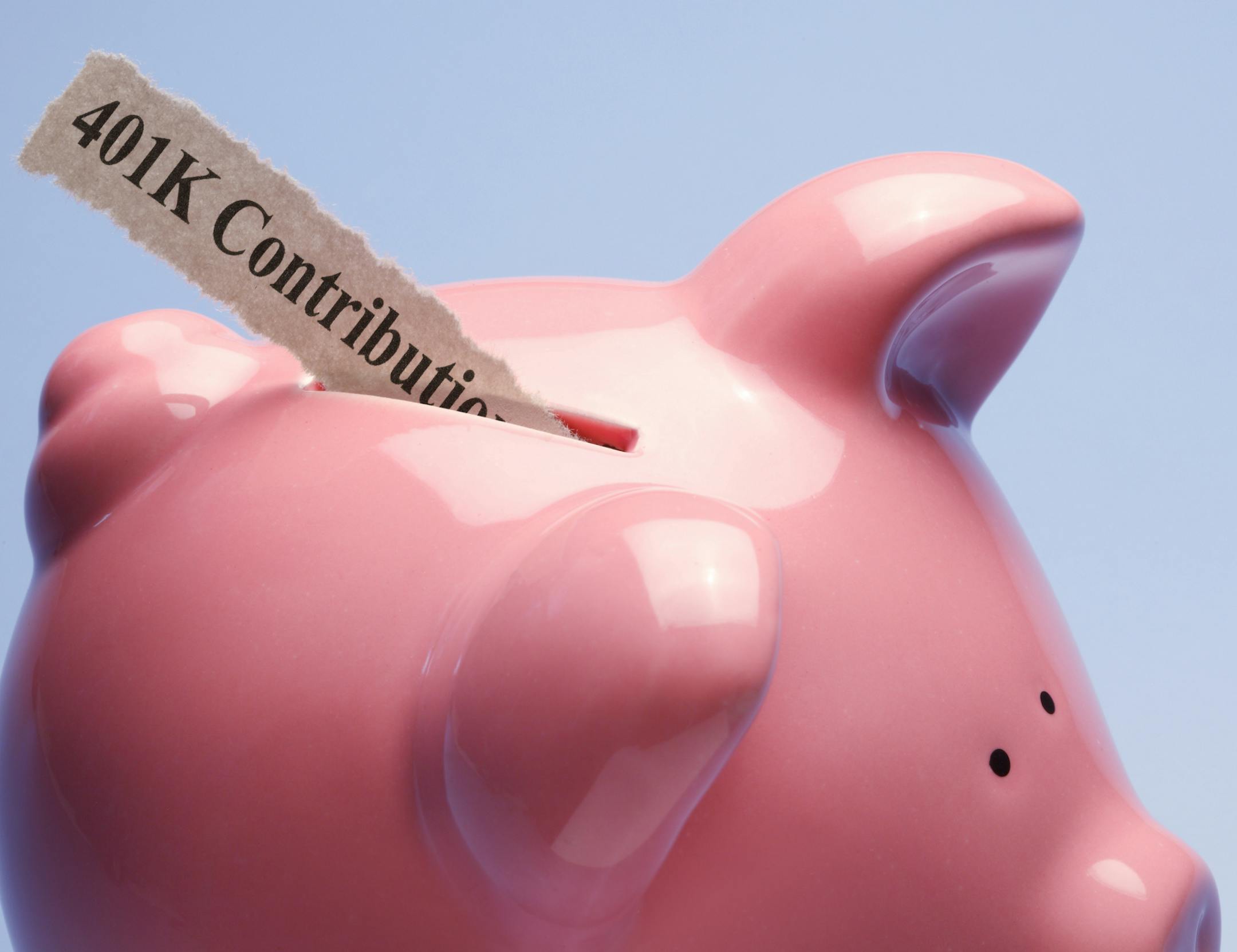 "401K Contribution" printed on a torn piece of paper that is inserted into the coin slot of a pink piggy bank against a blue background.