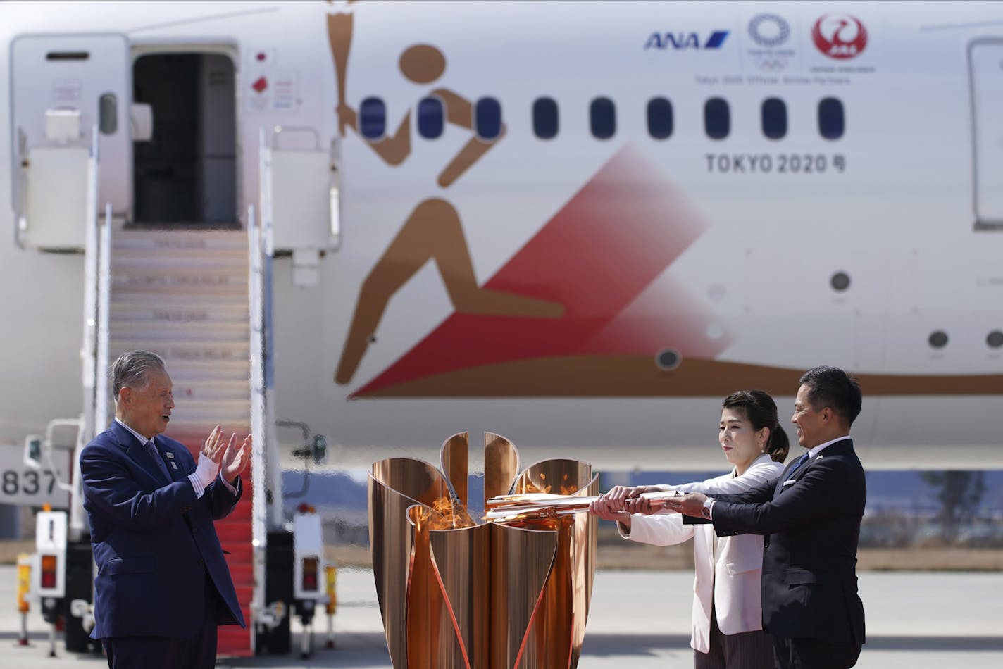 Three-time Olympic gold medalists Tadahiro Nomura, right, and Saori Yoshida light the torch as Tokyo 2020 Olympics chief Yoshiro Mori, left, watches during Olympic Flame Arrival Ceremony at Japan Air Self-Defense Force Matsushima Base in Higashimatsushima in Miyagi Prefecture, north of Tokyo, Friday, March 20, 2020. The Olympic flame from Greece arrived in Japan even as the opening of the the Tokyo Games in four months is in doubt with more voices suggesting the games should to be postponed or c