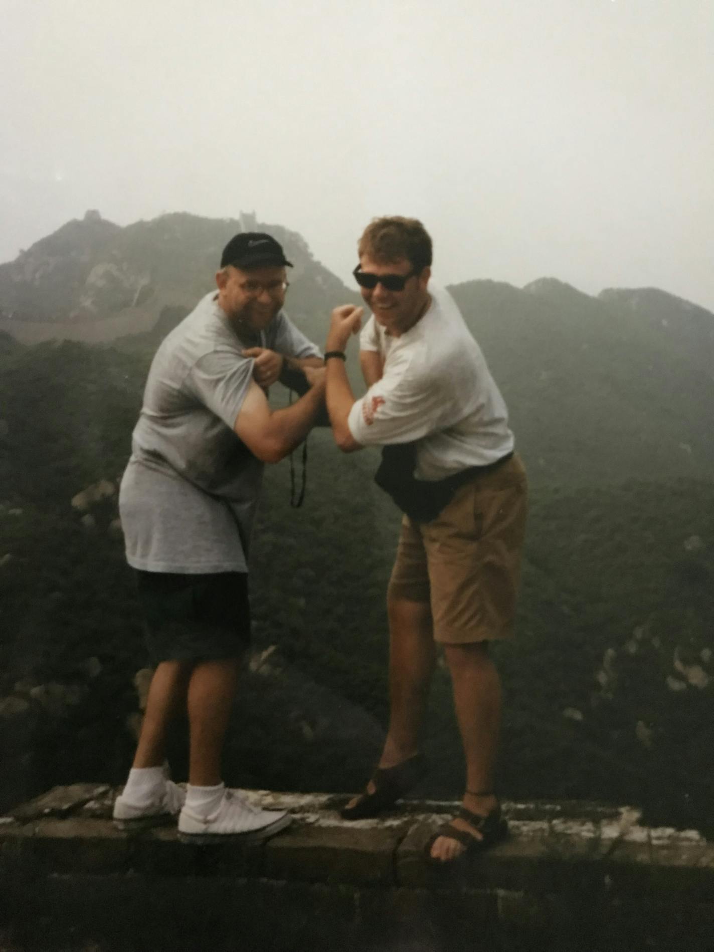 Two men flex their biceps and smile at the camera while standing on a ledge with mountains in the background.