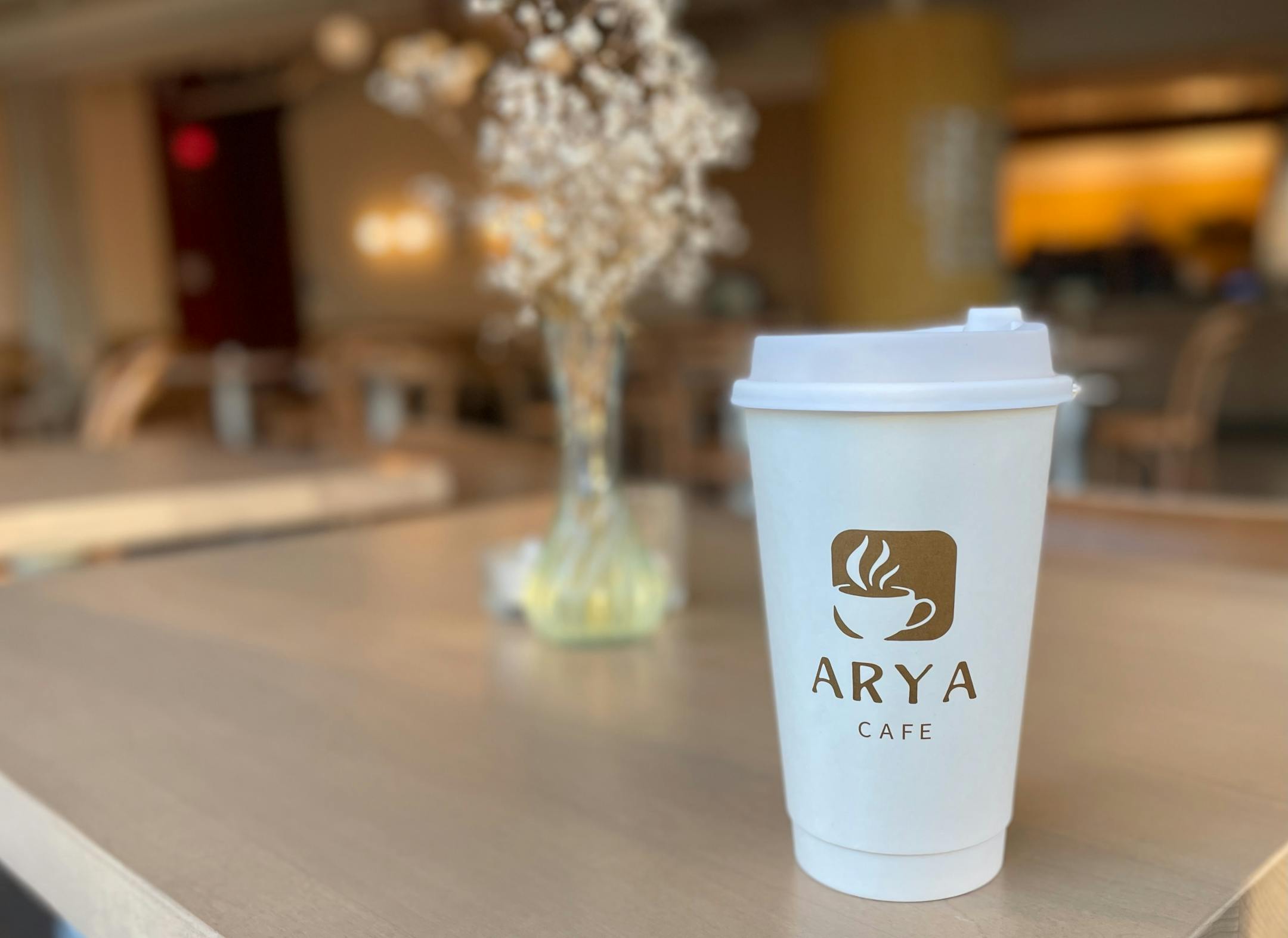 A latte in a branded white cup from Arya Cafe sits on a table decorated with a vase of baby's breath. The warm tones and light wood interior of the cafe is blurred in the background.
