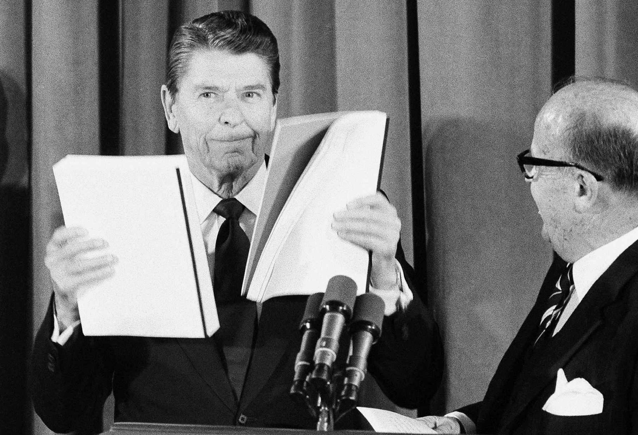 FILE - In this Jan. 16, 1984, file photo, President Ronald Reagan displays reports gives to him by J. Peter Grace, right, of the Private Sector Survey on Waste and Cost Control in Government, in Washington at the White House. The Grace Commission was created in 1982 by Reagan to root out waste and inefficiency in the federal government. Headed by Grace, the commission produced hefty recommendations it claimed would save the government $424 billion over three years. Reagan and Congress largely ig