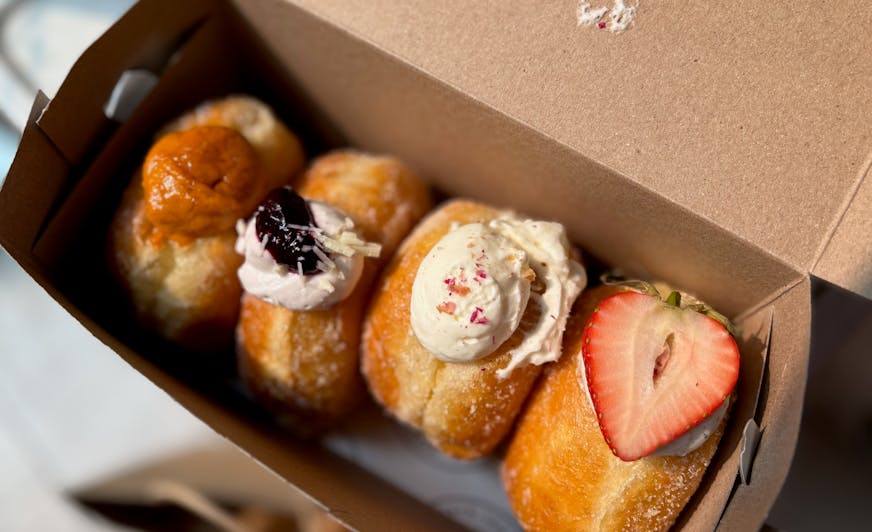 Four brioche stuffed doughnuts in a box with Thai tea, blackberry, rose lychee and strawberry black sesame flavors.