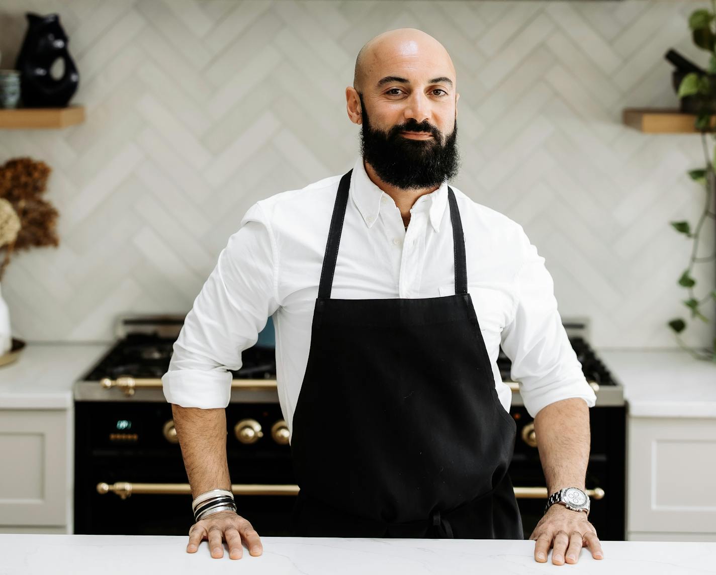 Chef Sameh Wadi in a white button down shirt and black apron in front of a white tiled background and black stove.