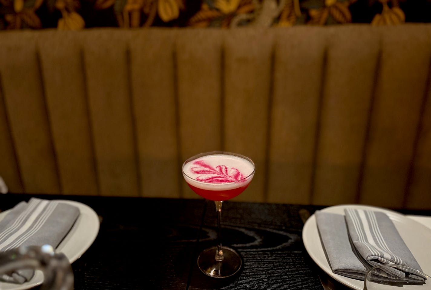 A wide shot of an empty, set table with a tall coup glass with a deep pink cocktail topped with white foam and garnished with a pink frond detail in the foam. The plush banquette and richly designed wallpaper are visible in the background.