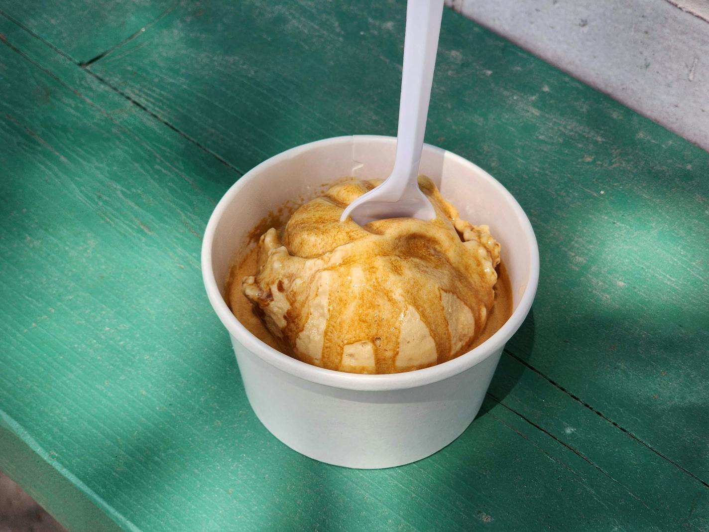 A green counter with a white cup containing a scoop of light brown ice cream with darker brown streaks of espresso running down. A spoon is stuck into the scoop.