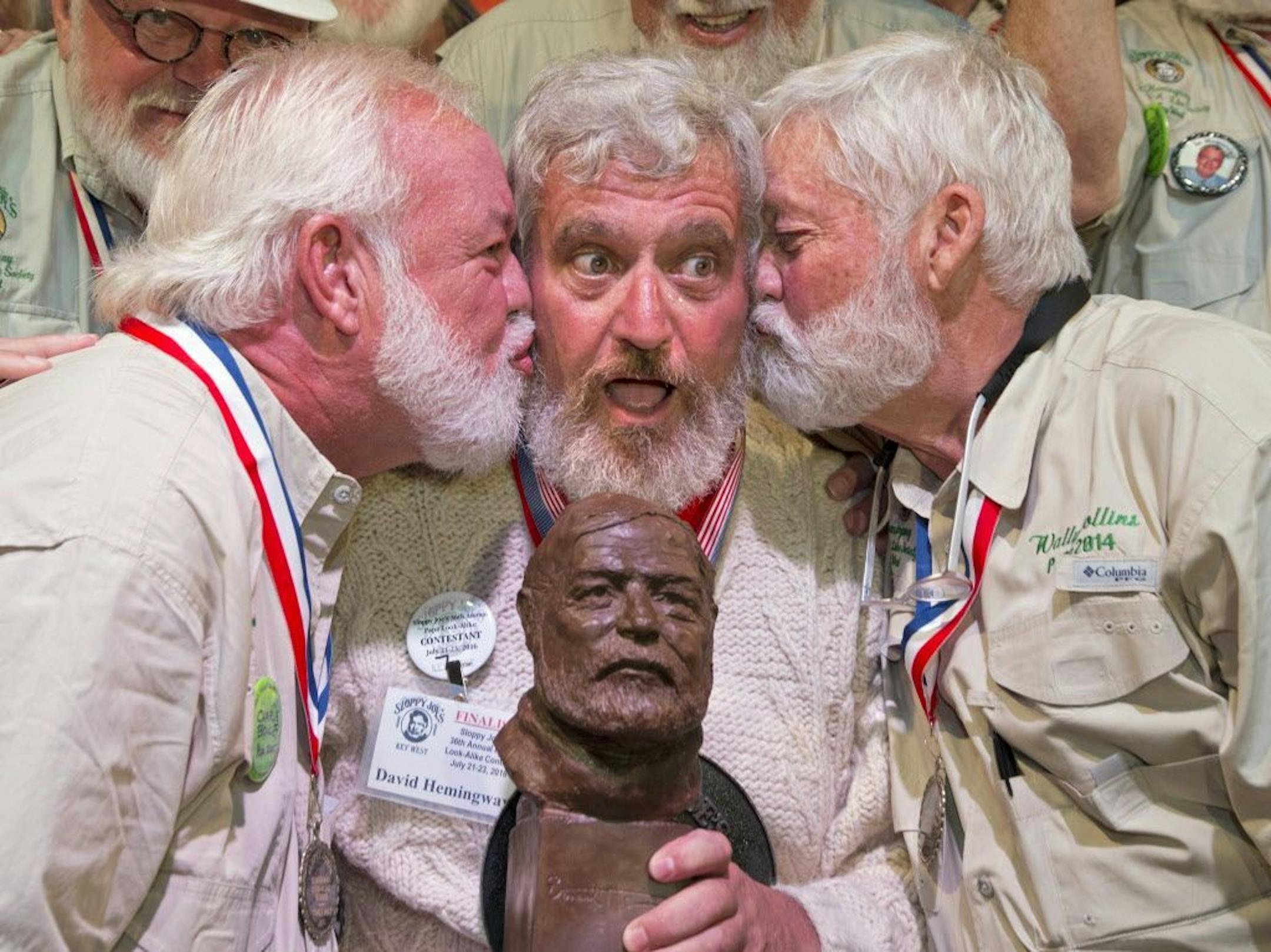 In this Saturday, July 23, 2016 photo provided by the Florida Keys News Bureau, Dave Hemingway, center, receives smooches from Charlie Boise, left, and Wally Collins, right, after Hemingway won the 2016 Ernest "Papa" Hemingway Look-Alike Contest in Key West, Fla. Success for Dave Hemingway came on the Macon, N.C., resident's seventh attempt to win the coveted title at Sloppy Joe's Bar. Dave Hemingway is not related to Ernest Hemingway who lived and wrote in Key West in the 1930s.