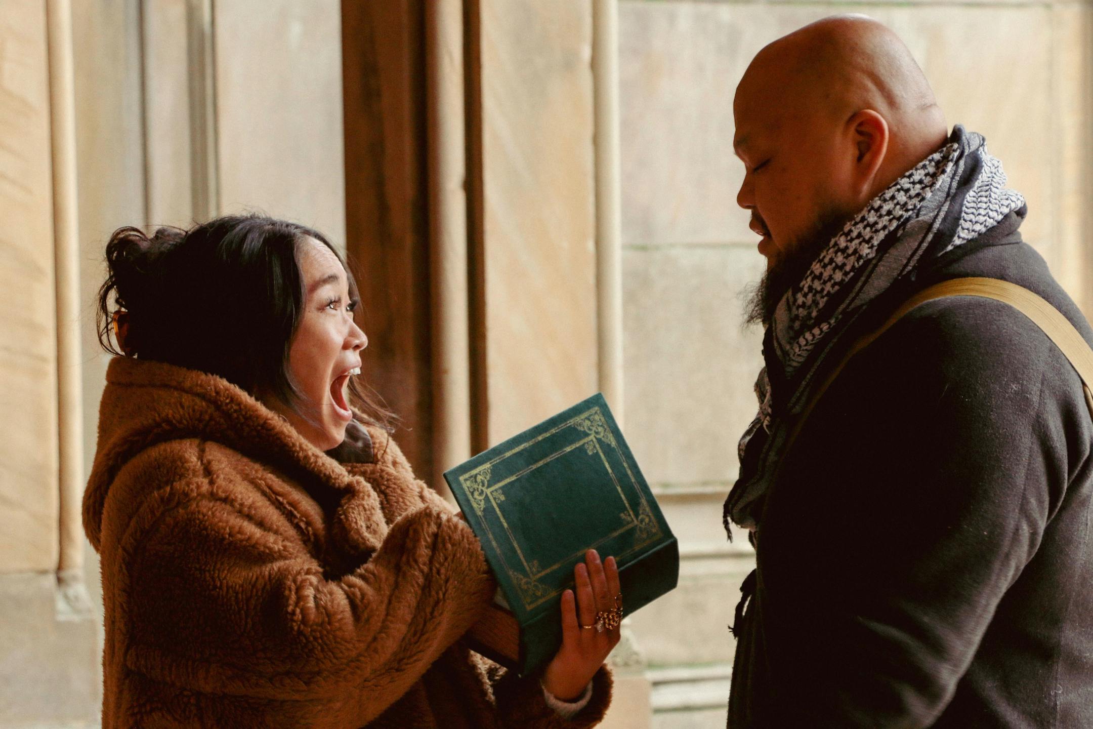 A woman, holding a fake book with something inside, opens her mouth in shock while looking at the man who gave it to her.