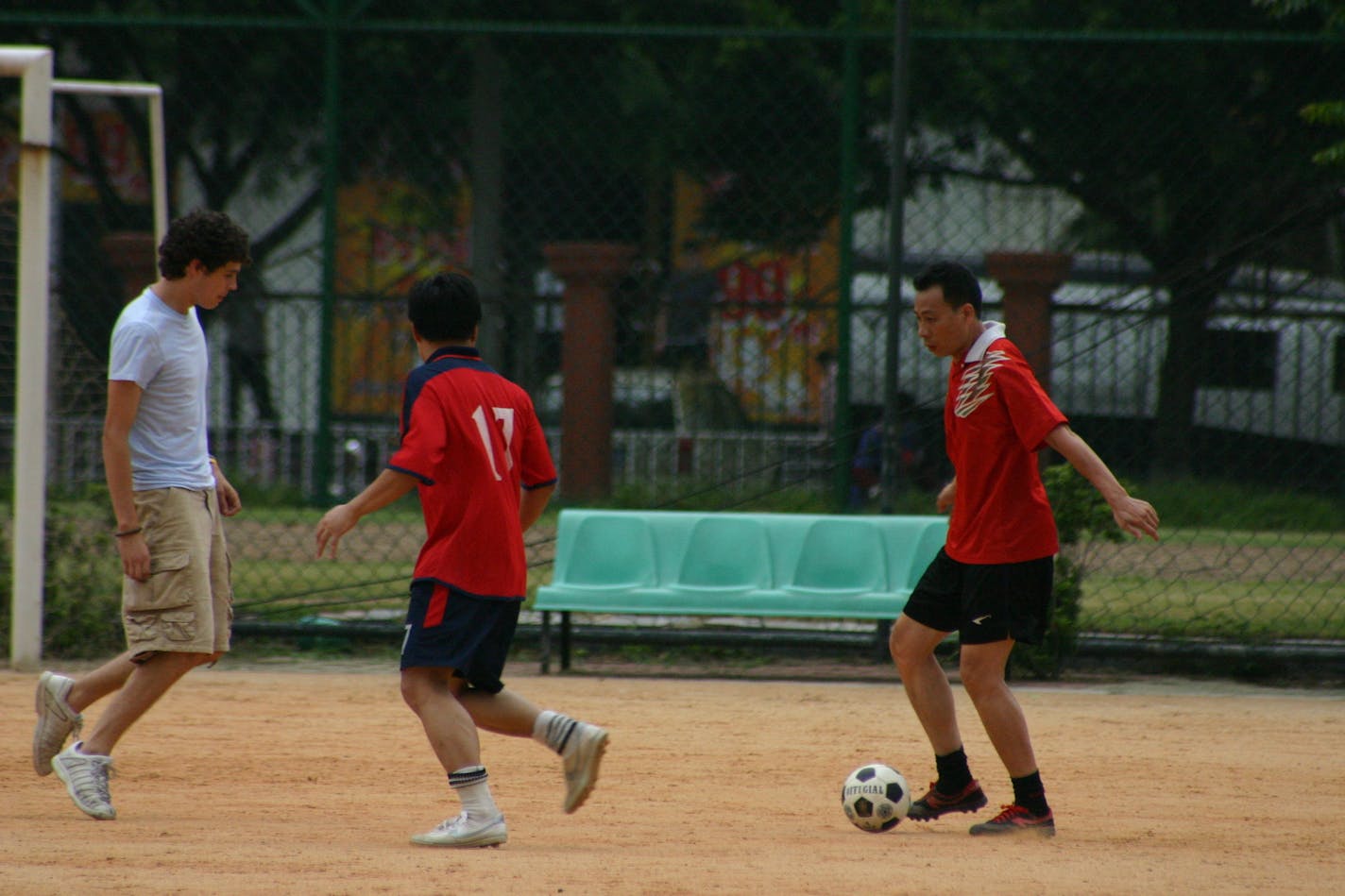 Two Chinese young men in red play soccer on a dirt pitch with a white high school student.