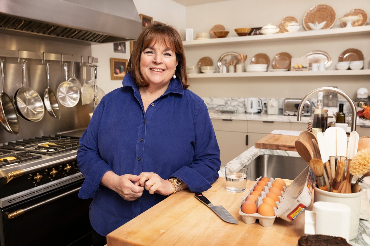 Ina Garten at her kitchen island, wearing a blue shirt, with eggs and knife on the surface and shelving and cabinets in the background with plates and appliances, and the stove to the left with pans hanging over it.