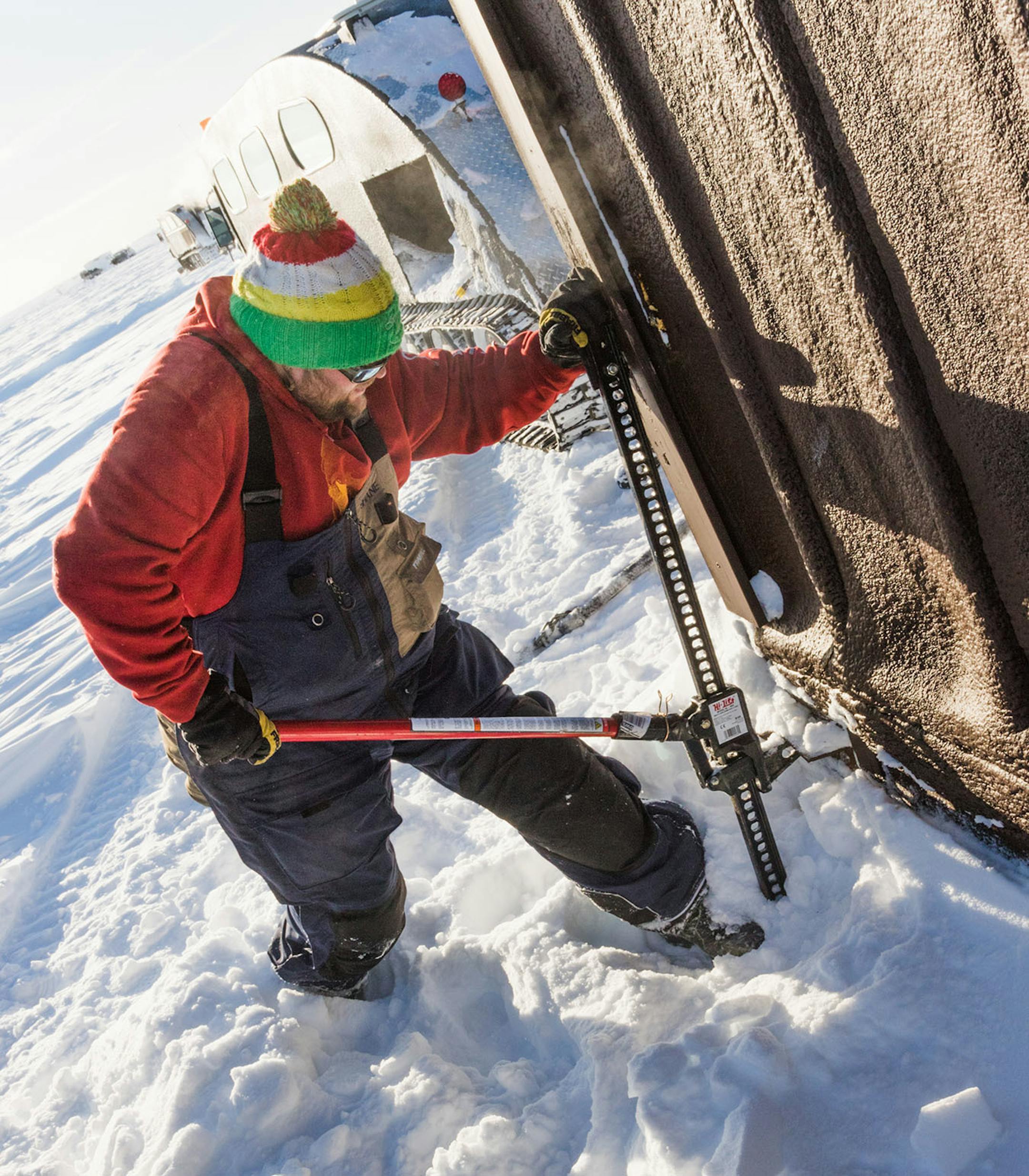 Shane Burk, an ice captain, uses a jack to lift an ice house up so he can attach chains to each side. The chains allow the guides to drag the ice houses with a bombardier to different locations.