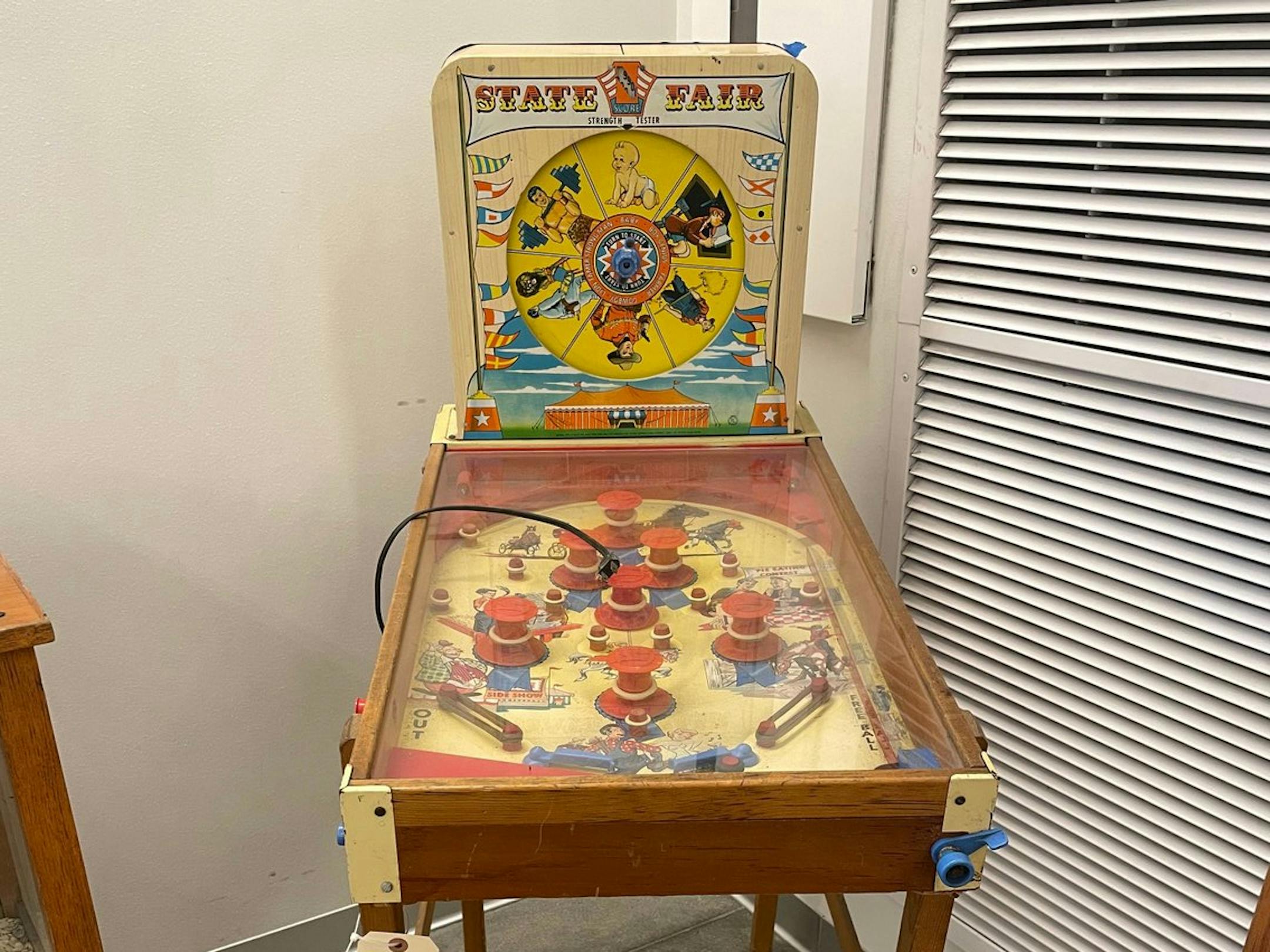 A small, ancient pinball machine with the words "State Fair" emblazoned across the top and a spinning wheel illustrated with pictures of a baby, a weightlifter, a cowboy, a farmer and two other figures.