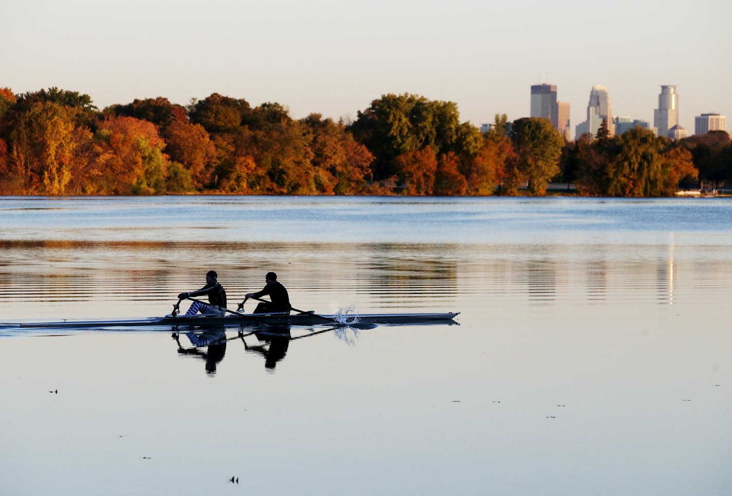 A rowing crew skims across a placid Lake Nokomis with fall colors and the Minneapolis skyline.