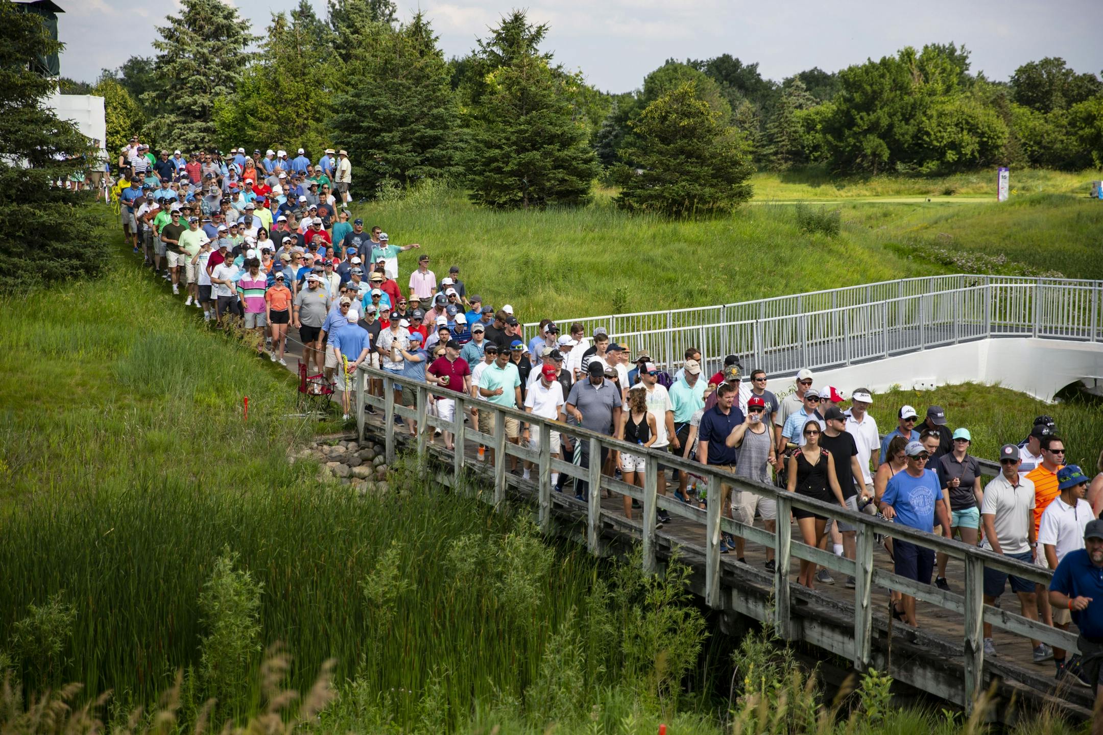 A large crowd of people move across the bridge from the 17 tee to the green.