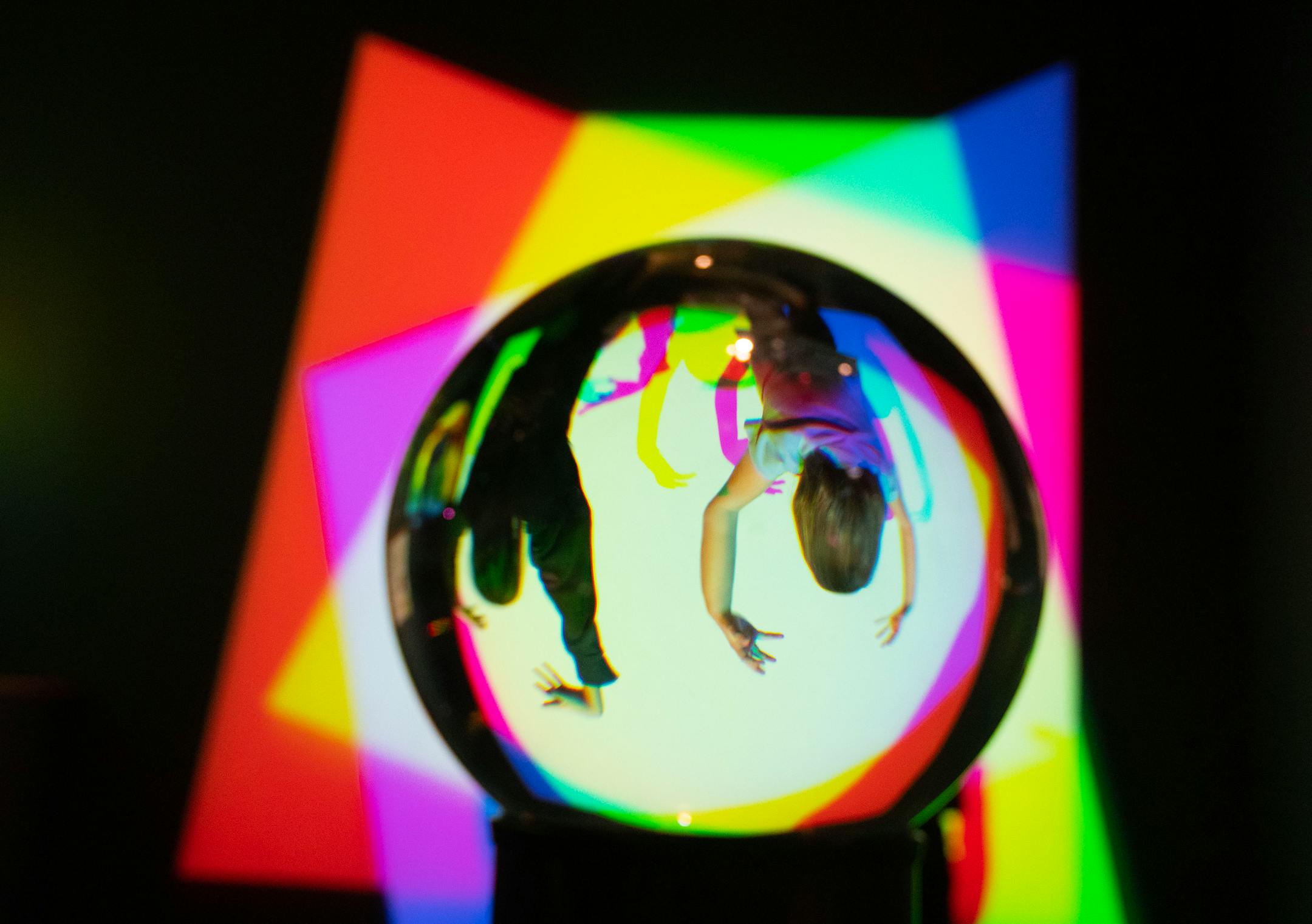 Children are reflected in a crystal ball as they play in an exhibit with colorful lights and shadows.