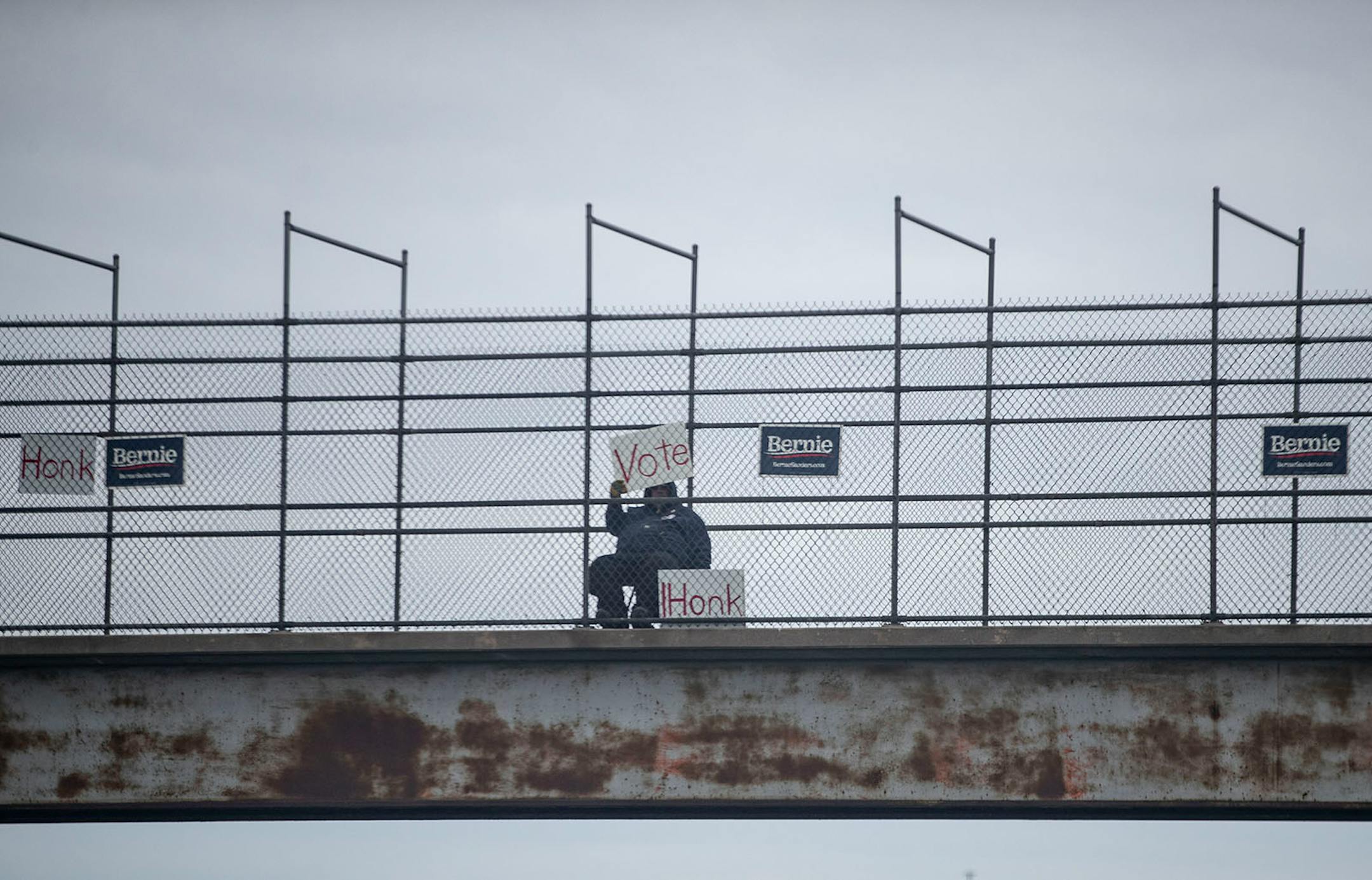 A man sat on a footbridge over Interstate 35 in Duluth on Tuesday morning, holding a sign encouraging people to vote for Bernie Sanders, with Sanders posters surrounding him.