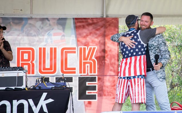 Mike Waldron hugs a man in a stars and stripes outfit while standing on a stage.