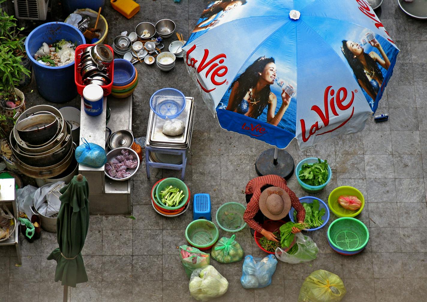 Under an umbrella advertising French bottled water, a woman prepares vegetables. Some of the cuisine of Vietnam, such as baguette sandwiches, has a French influence.