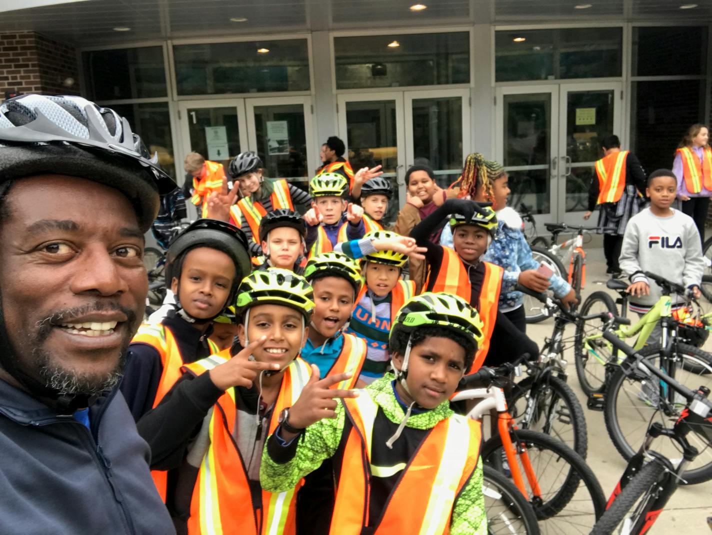 A Black man wearing a helmet and windbreaker smiles while a dozen or more racially diverse kids in orange vests and helmets pose for the camera alongside several bikes.