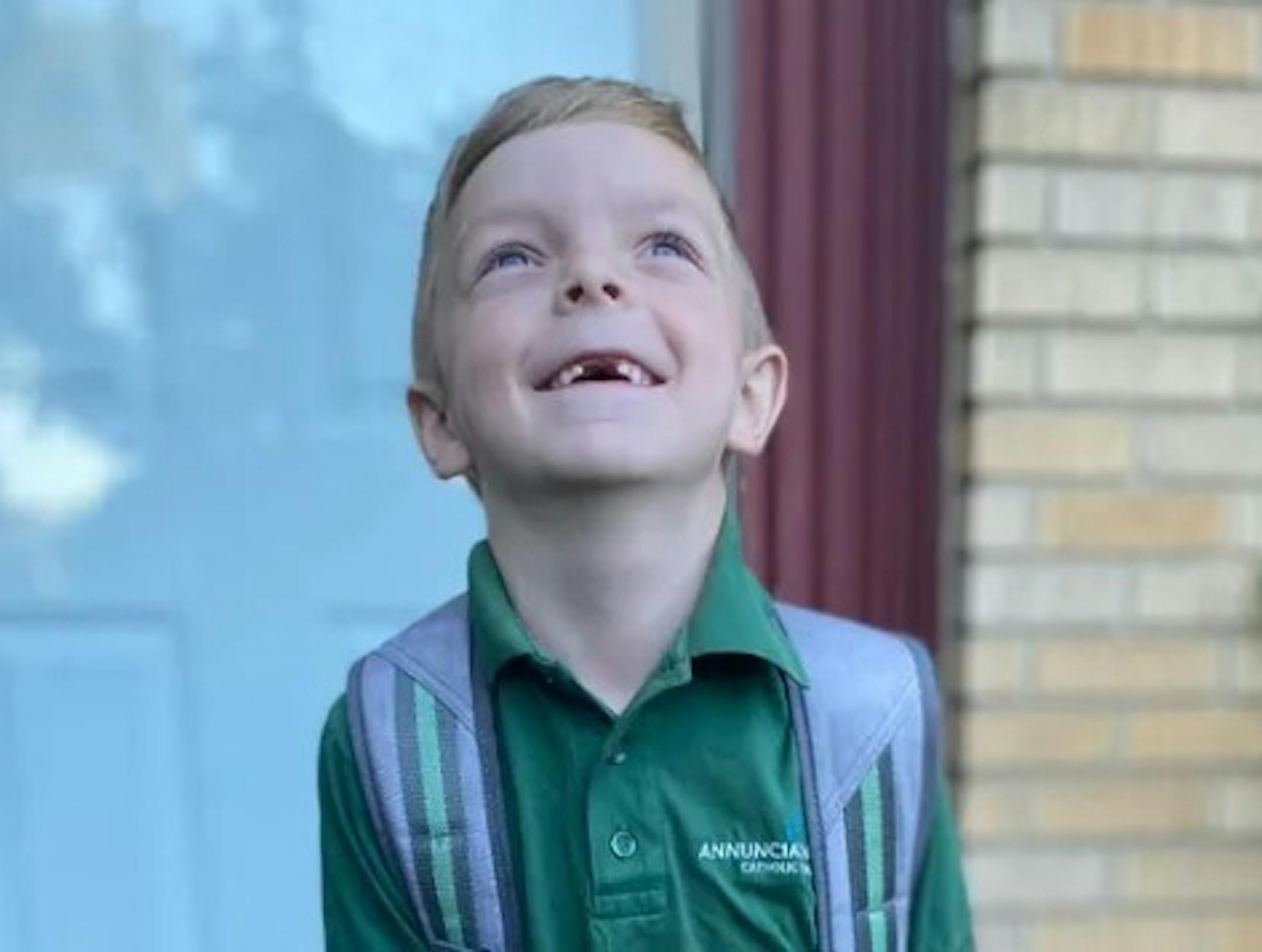 A smiling boy with two missing front teeth wears a backpack and a green school uniform polo smiles on what appears to be his front stoop.