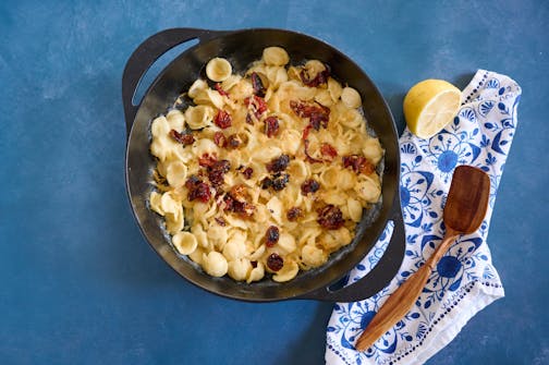 A pan with cheesy pasta and roasted cherry tomatoes next to a wooden spoon and bright napkin.