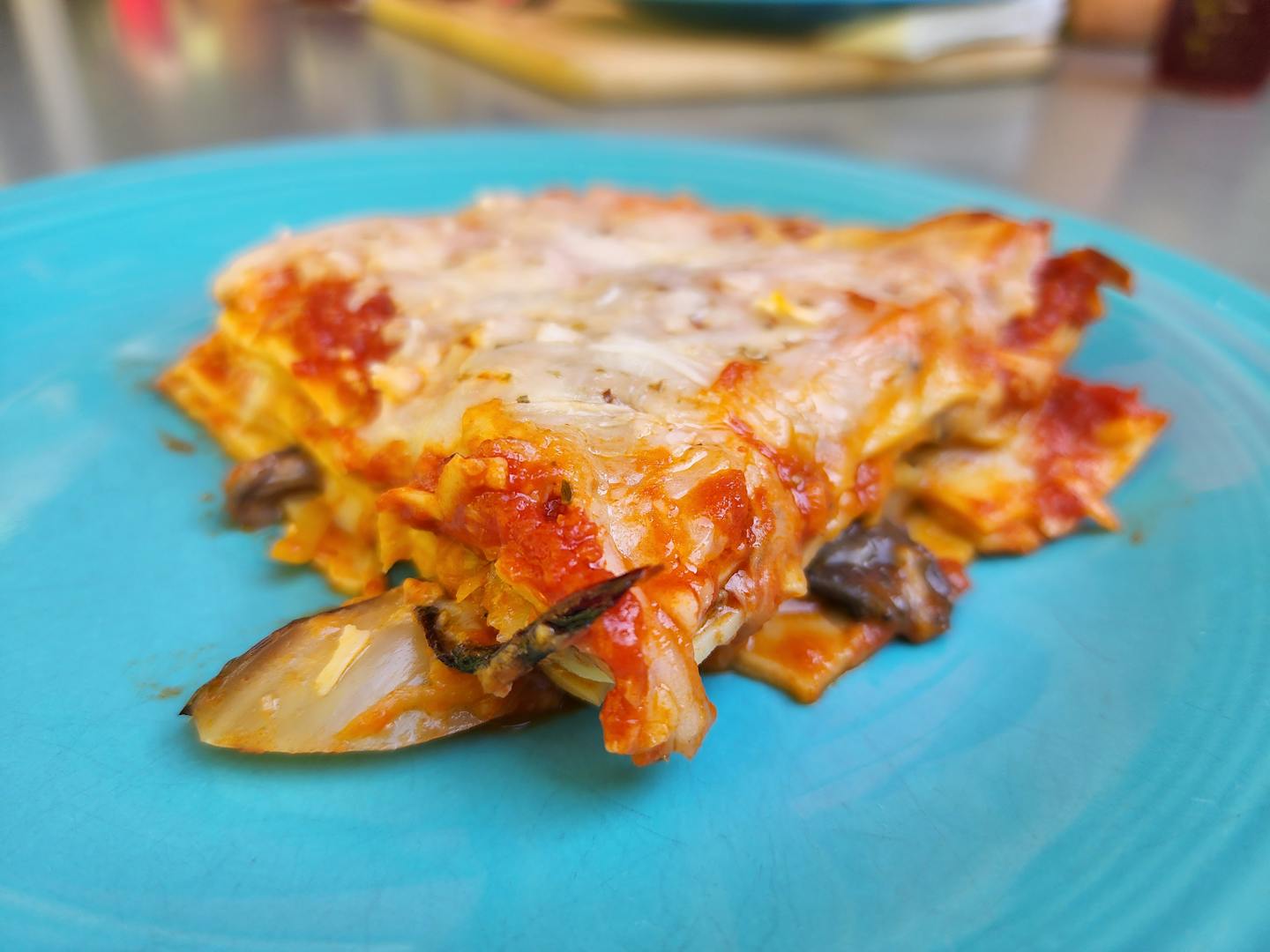 A square of lasagna, red sauce and vegetables coming out the sides and melted cheese on top, served on a blue plate.