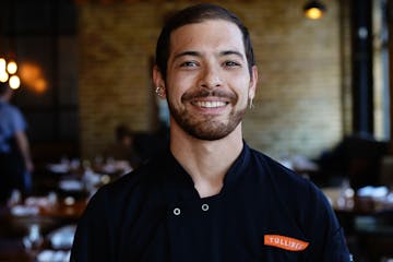 The smiling chef with hair tied back stands in the Tullibee dining room.