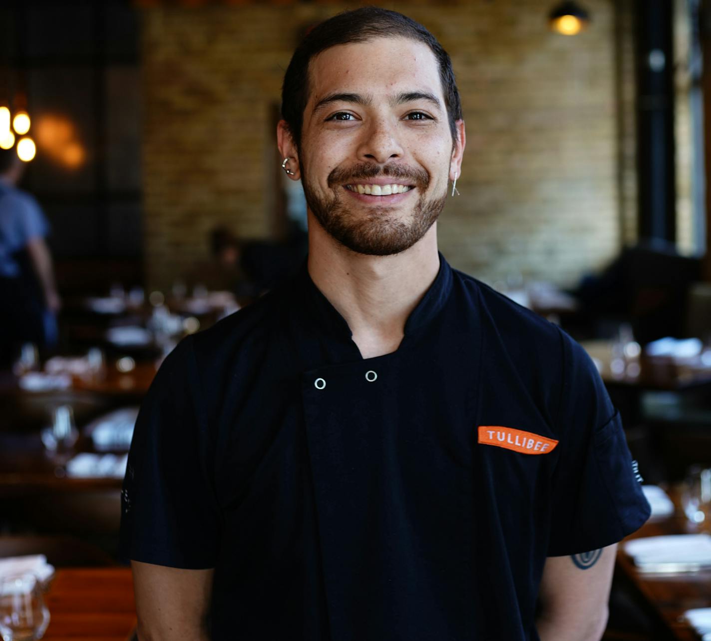 The smiling chef with hair tied back stands in the Tullibee dining room.