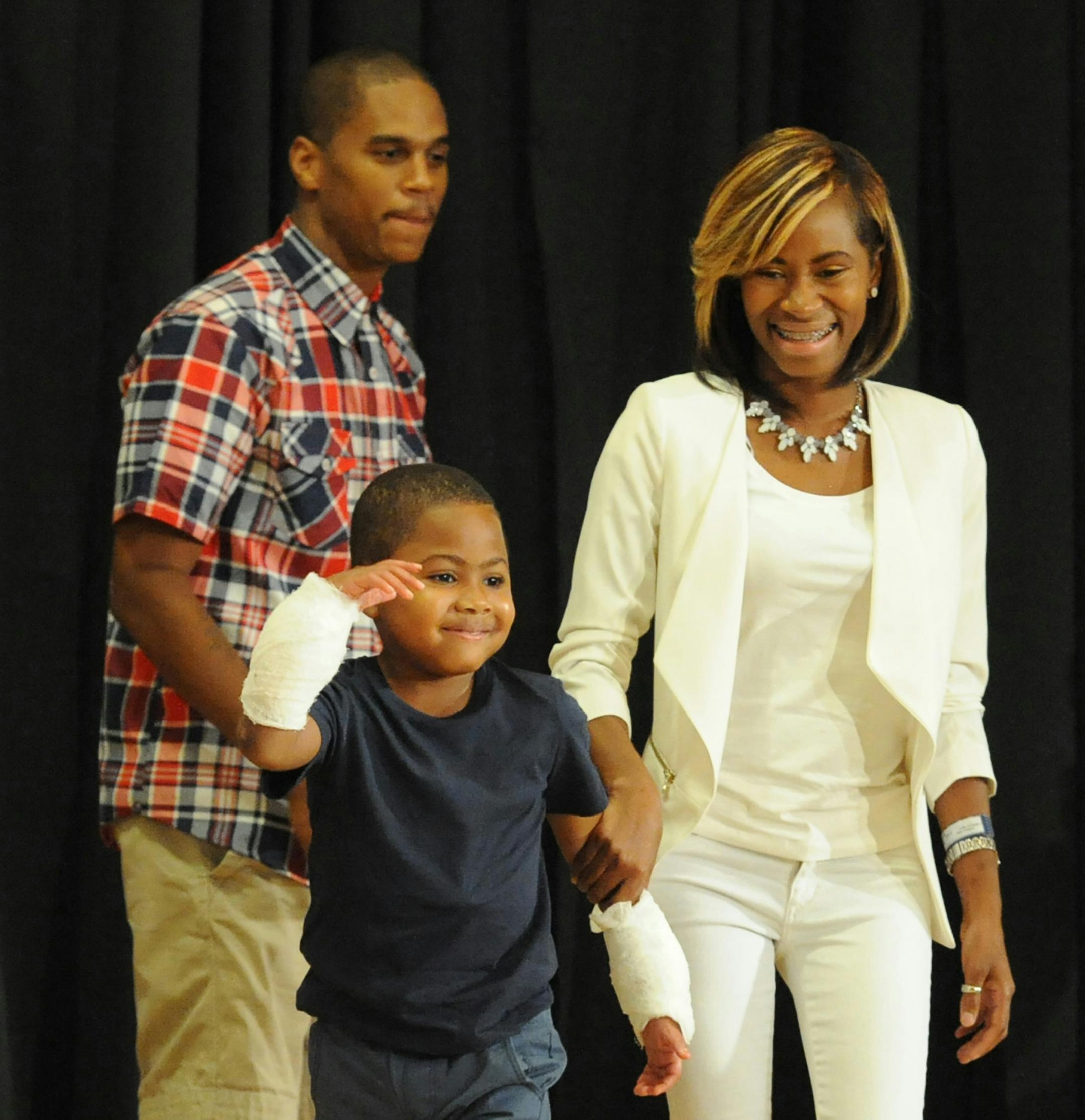 Zion Harvey, 8, of Baltimore, waves to the audience with his new right hand as his mother, Pattie Ray, leads him onstage to the at a news conference at Children's Hospital of Philadelphia on Tuesday, July 28, 2015. Zion, who lost his hands and feet to a bacterial disease as a two-year-old, had a double hand transplant at CHOP in early July 2015, the first pediatric double hand transplant. In the background is his step-father Kevon Gant. (Clem Murray/Philadelphia Inquirer/TNS)