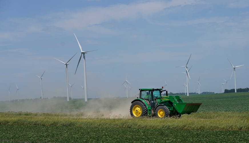A farmer drives his tractor along a gravel road, below wind turbines from the Fenton Wind Farm, where power line congestion is increasingly shutting down and leading to significant tax revenue decline Wednesday, June 29, 2022 in Murray County, Minn.