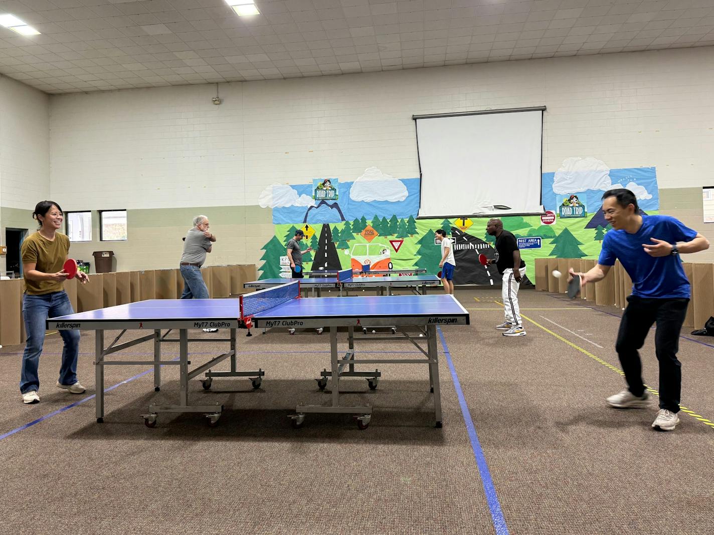Two people smile as they play table tennis in a church basement filled with ping pong tables.