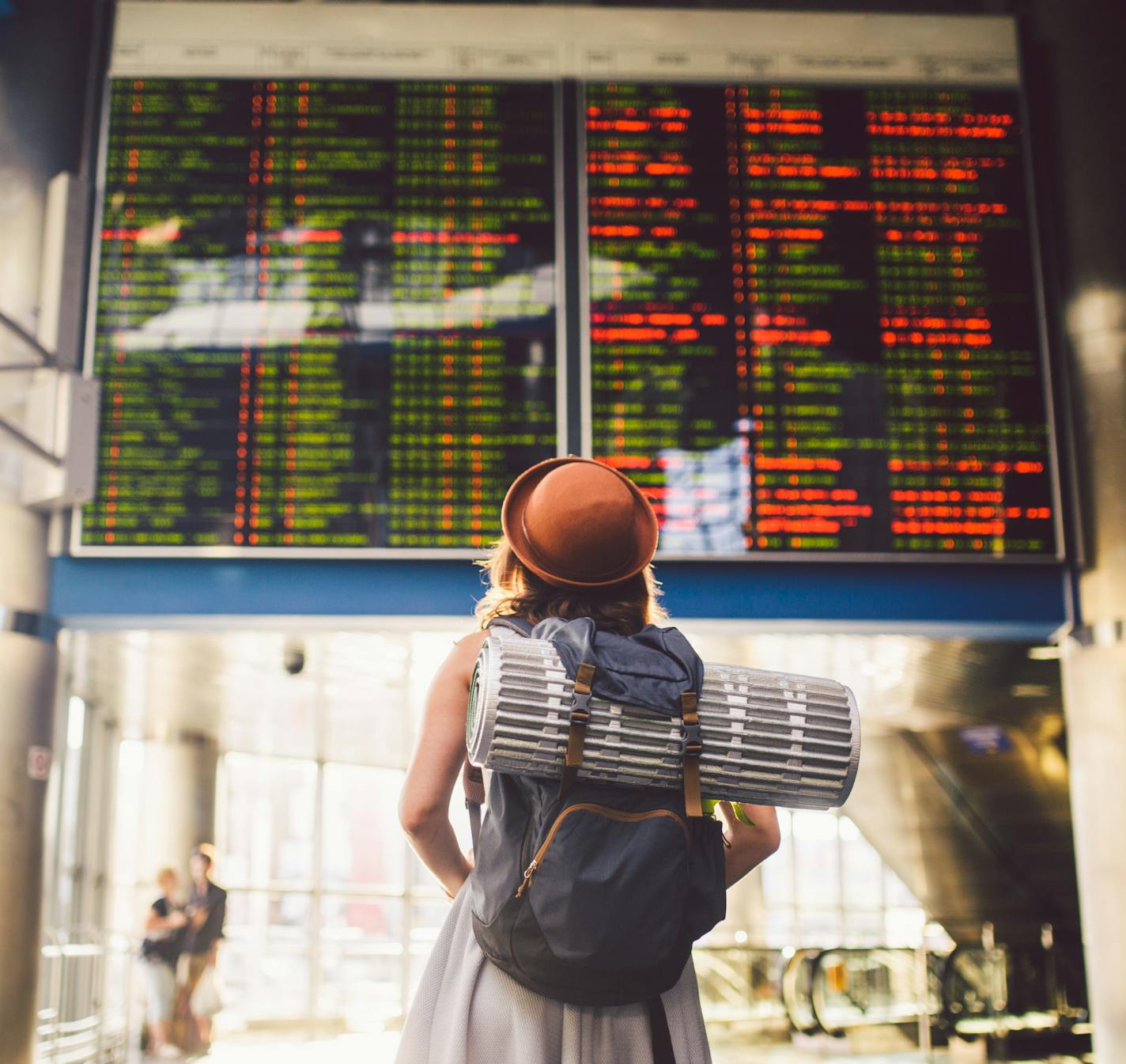 Theme travel public transport. young woman standing with back in dress and hat behind backpack and camping equipment for sleeping, insulating mat looks schedule on scoreboard airport station sunny day