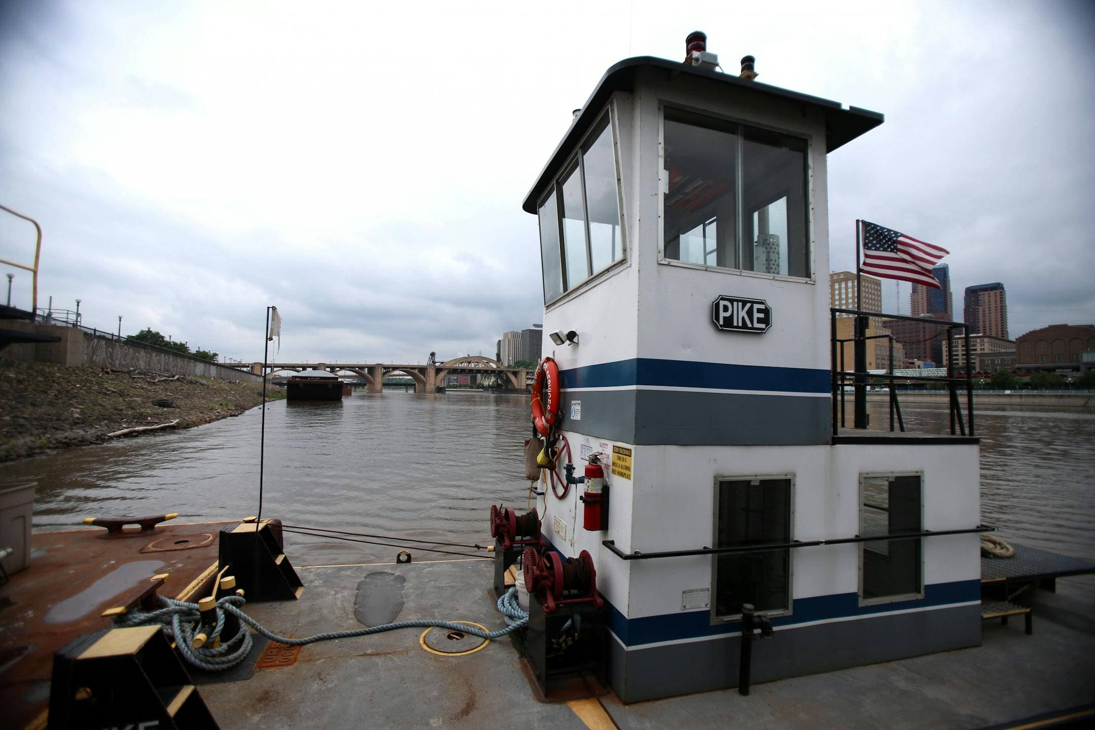 The Pike is the tug boat the five men appropriated in order to rescue the individual from the Mississippi River. In the background, the bridge that the individual jumped from, the Roberts Street Bridge, can be seen.